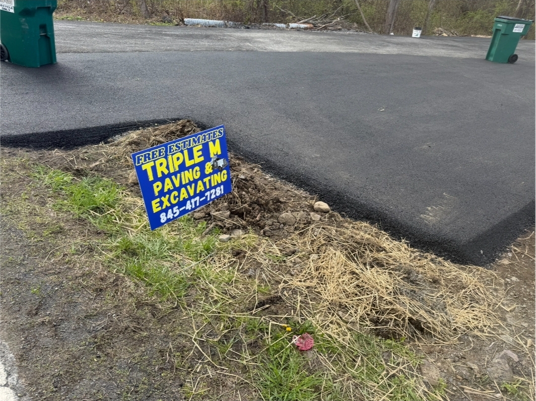 Freshly paved driveway with a small patch of exposed soil and grass. A yellow and blue sign reads Triple M Paving & Excavating with a phone number for free estimates.