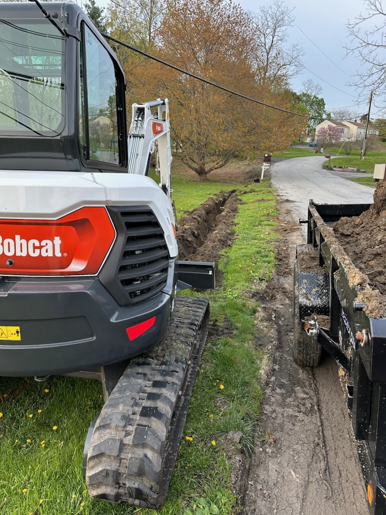 A small excavator sits on grass next to a trailer filled with dirt, with a freshly dug trench running alongside a suburban street.