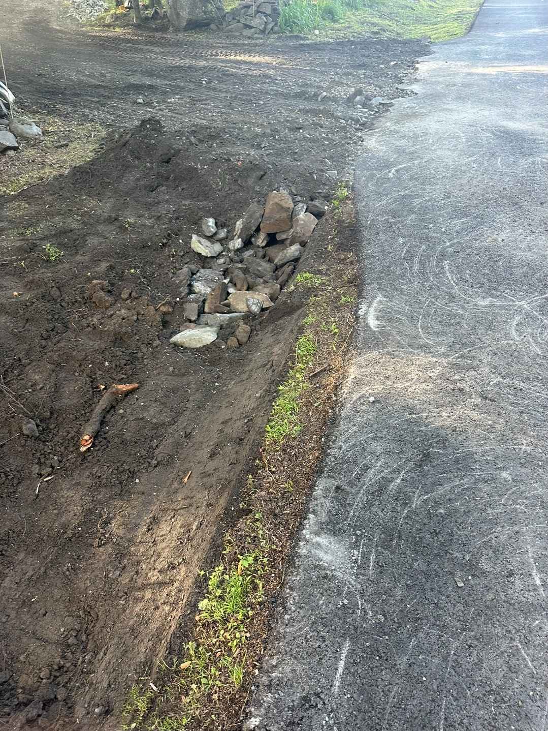 Dirt ditch lined with rocks runs alongside a paved road; grass and soil are visible at the edge of the pavement.