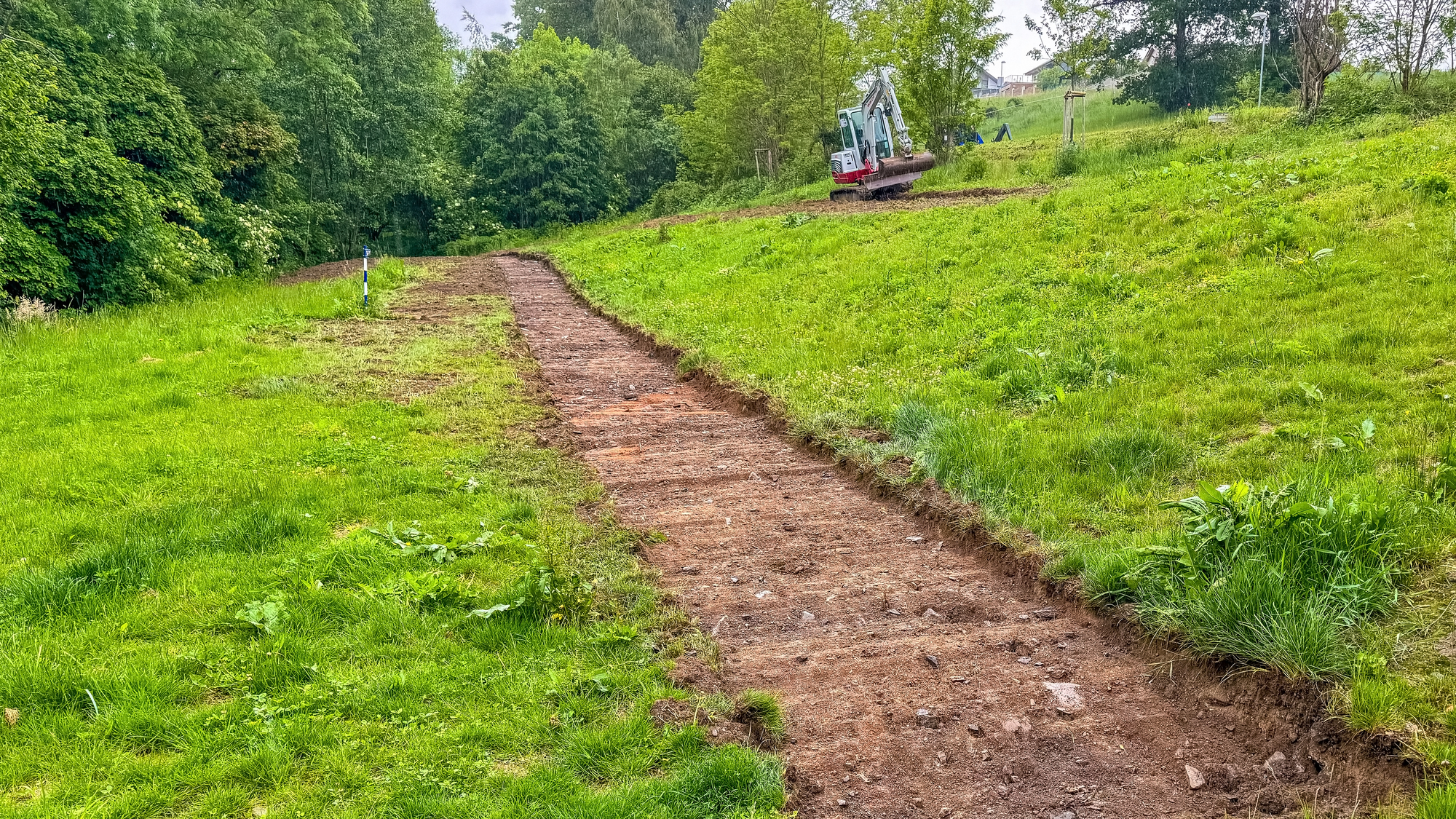 A narrow dirt path has been freshly cleared through grassy terrain, leading toward an excavator positioned on a gentle hill in the distance.
