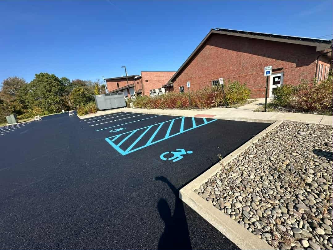 Freshly paved parking lot with two marked accessible parking spaces and a blue-striped access aisle next to a brick building under a clear sky.