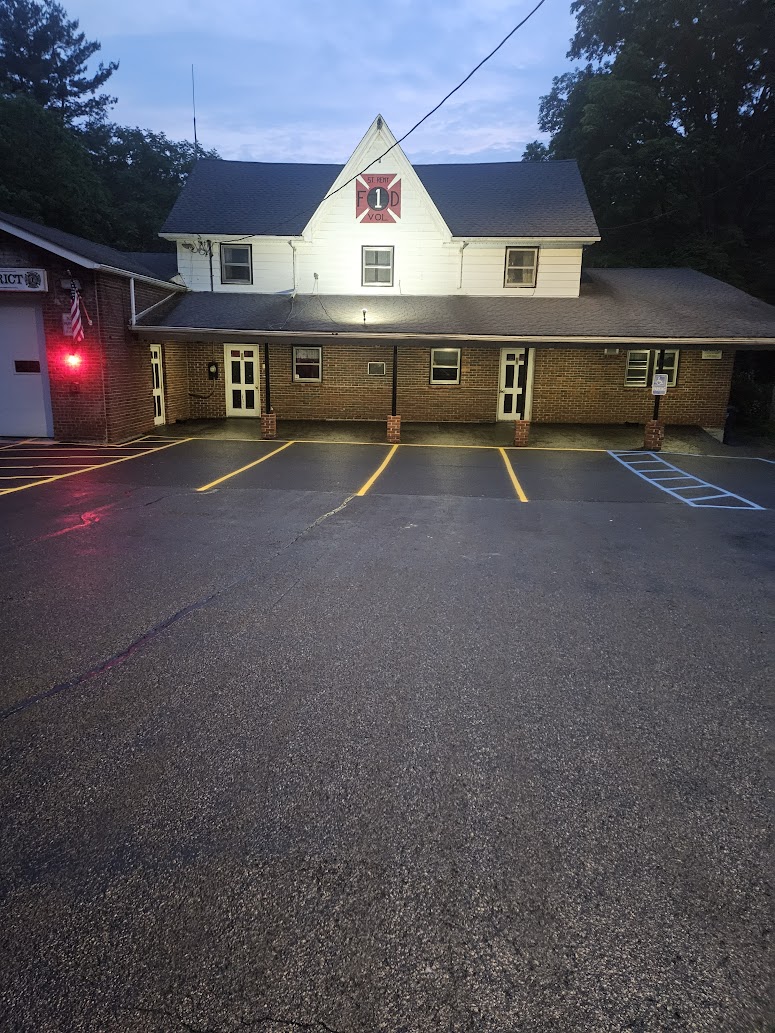A brick fire station building with a red fire department emblem above the entrance, illuminated at dusk, with empty, freshly painted parking spaces in front.