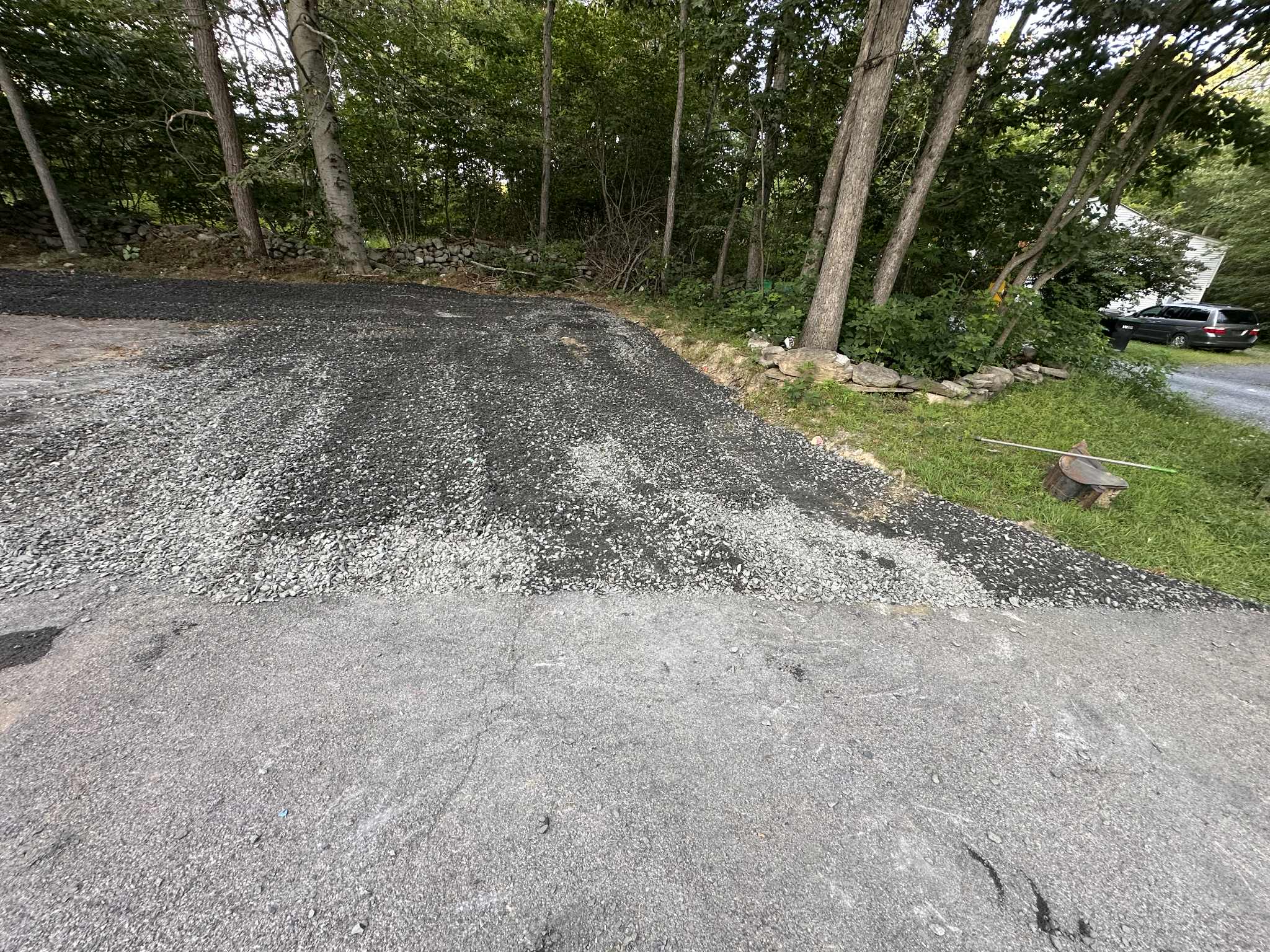 A driveway is partially covered with new gravel beside a wooded area, with a car and garden tools visible in the background.