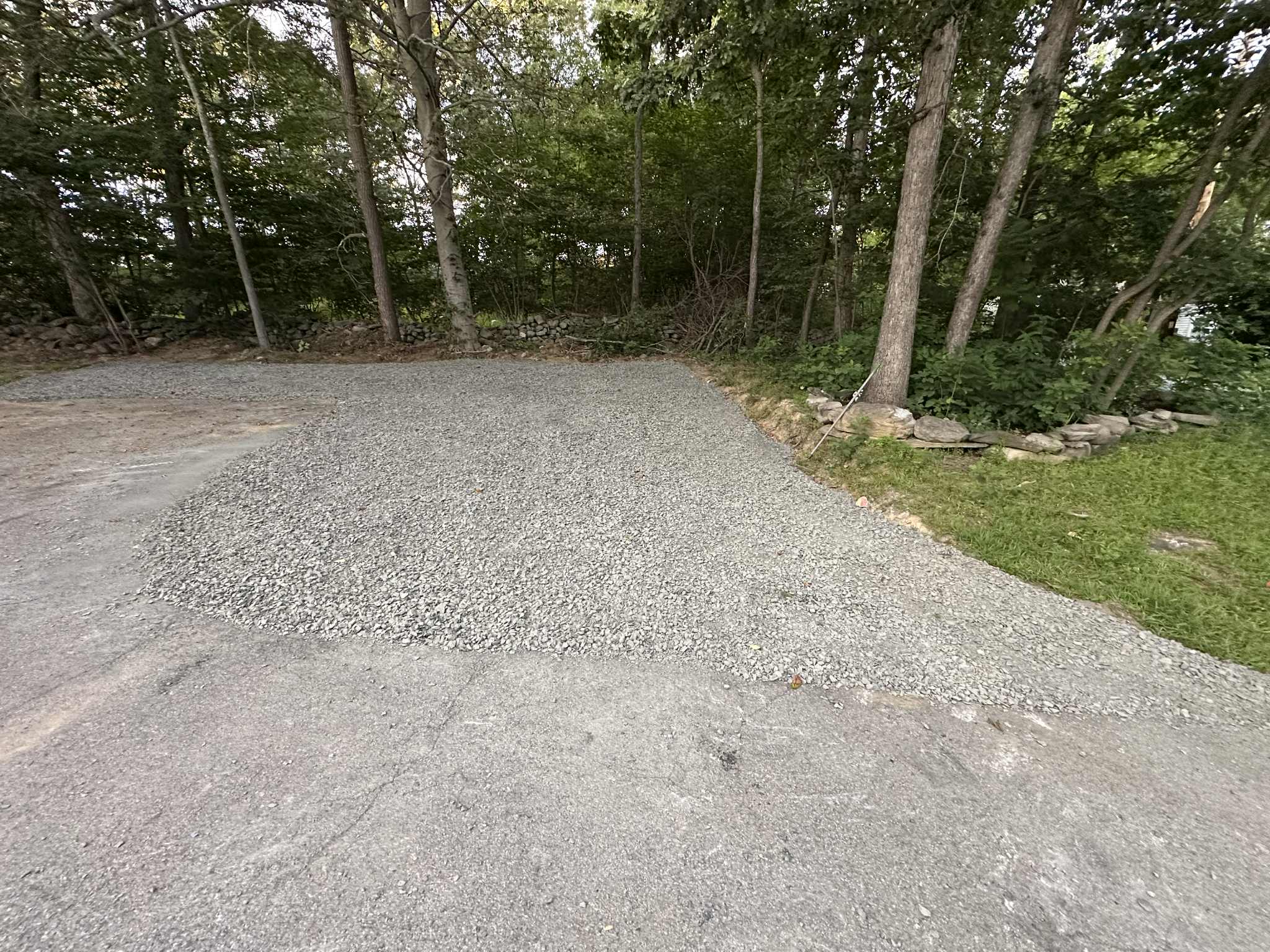 A section of gravel has been spread over a partially paved area next to a patch of grass and trees in a wooded outdoor setting.
